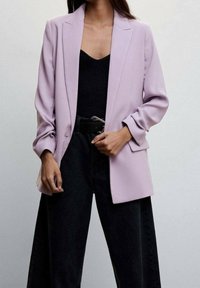 Blazer lavande à coupe ajustée, avec des revers crantés et des manches amples, porté sur un haut noir et associé à un pantalon large noir.
