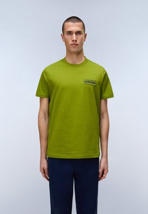 Uomo con capelli corti che indossa una maglietta verde lime con piccolo logo sul petto e pantaloni blu scuro, in piedi davanti a uno sfondo chiaro e uniforme.