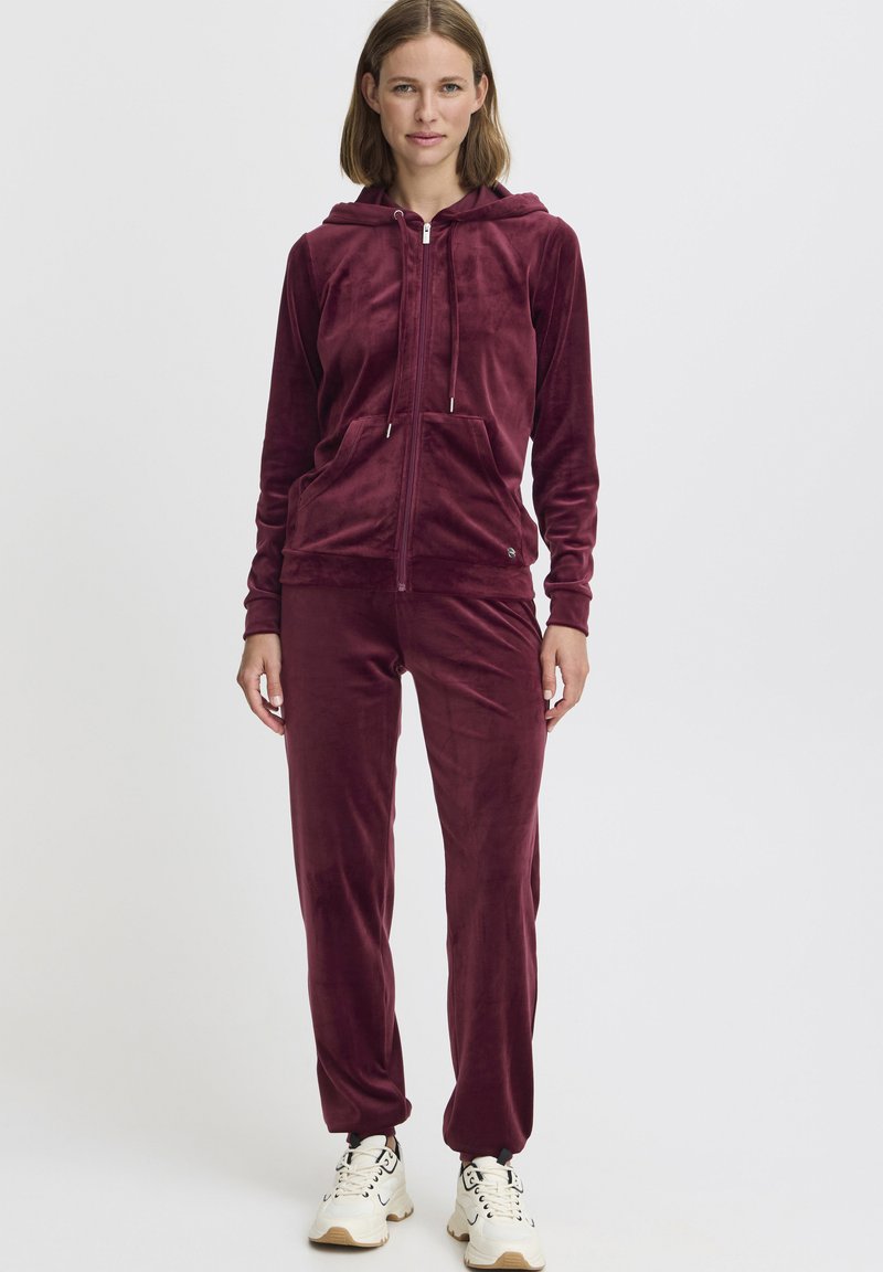Survêtement en velours bordeaux avec une veste à zip et des poches avant ; pantalons ajustés à jambe droite. Assorti avec des baskets blanches imposantes.