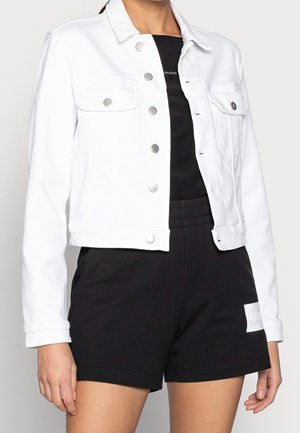 Vrouw draagt een witte spijkerjas met knopen over een zwart topje en zwarte shorts met elastische taille en een wit patch op het linkerbeen.