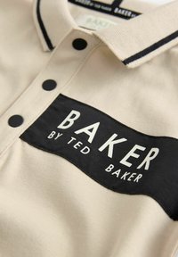 Bež polo majica s crnim zakrpom na kojoj je napisano "BAKER" bijelim slovima, crnim gumbima i kontrastnim ovratnikom s detaljima šivanja.