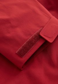 Röd tyg med en slät yta, som har en kardborreknäppning vid manschetten. Enkelt mönster med en vikning och sydda kanter.