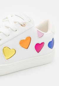 Białe trampki z kolorowymi łatkami w kształcie serca w kolorach żółtym, pomarańczowym, różowym i niebieskim. Posiadają płaskie sznurowadła oraz teksturowaną gumową podeszwę.