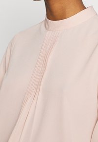 Blouse en tissu rose doux avec des détails plissés sur le devant, dotée d'un col montant et d'une texture lisse.