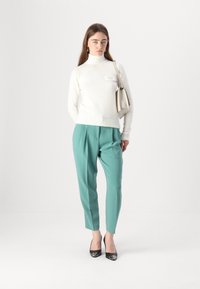 Maglione bianco a collo alto, pantaloni teal, tacchi neri alti con dettagli metallici. La modella tiene una piccola borsa beige. Sfondo bianco uniforme.