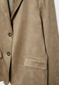 Gros plan d’un blazer en daim beige avec revers, deux boutons foncés et une poche à rabat sur le devant côté droit.