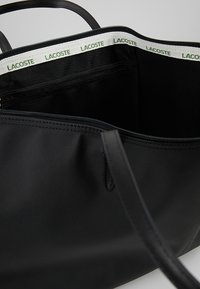 Černá taška přes rameno se zipem, s bílým vnitřním lemováním označeným "LACOSTE" a černými uchy.
