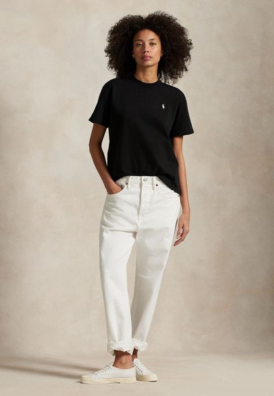 Femme aux cheveux bouclés debout, portant un t-shirt noir à manches courtes, un pantalon blanc taille haute et des baskets blanches, sur un fond uni.