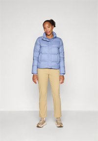 Lyseblå vattert jakke med knappelukking og høy krage. Brukt over beige bukser og joggesko, med en glatt tekstur og quiltet design.