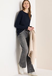 Granatowy sweter dzianinowy, szare rozszerzane dżinsy i białe trampki. Trzyma beżowy sweter z polaru. Gładka tkanina i luźny krój na całej powierzchni.