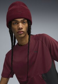 Maroon short-sleeve shirt with black accents and a subtle logo. Knitted maroon beanie with a textured pattern. Model has long braided hair.