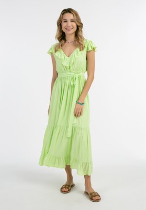 Rochie maxi de culoare verde deschis, cu mâneci volan, decolteu în V, talie strânsă și fustă stratificată. Material ușor, asociată cu sandale brodate.