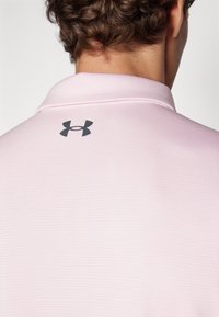 Camisa polo rosa con un diseño texturizado, que presenta un logo negro de Under Armour en el cuello de la parte posterior.
