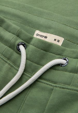 Zelené bavlněné joggingové šortky s nastavovacími bílými šňůrkami a štítkem s brandingem "ombre". Textura je hladká a měkká.