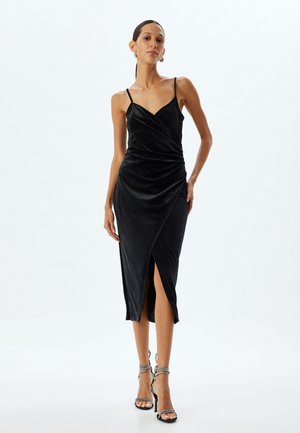 Schwarzes Samt-Midikleid mit Spaghettiträgern, Wickeldesign, seitlichem Schlitz und leichtem Glanz. Kombiniert mit silbernen Riemchen-Heels für einen Kontrast.