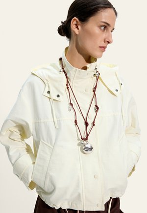 Młoda kobieta z włosami upiętymi w kok, ubrana w jasnosłoneczną żółtą kurtkę oraz nałożone na siebie czerwone sznureczki z srebrnymi wisiorkami, patrząca na bok.