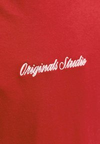 Fehér hímzett "Originals Studio" felirat piros szövet háttéren, ruházati részlet kiemelésével.