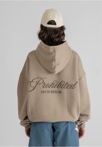 Overdimensjonert beige hettegenser med hette, med teksten "Prohibited 10119 BERLIN" i mørkegrønn skrift på ryggen. Laget av bomullsblandet stoff.