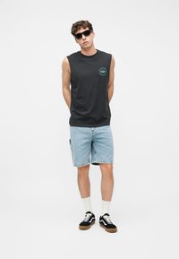 Giovane uomo con indosso una maglietta nera senza maniche, pantaloncini di jeans azzurri, calzini bianchi, scarpe da ginnastica nere e occhiali da sole neri, in piedi davanti a uno sfondo bianco.