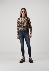 Mujer vestida con una blusa de manga larga de estampado de leopardo, jeans oscuros ajustados, sandalias negras gruesas, gafas de sol y llevando un bolso negro de hombro, frente a un fondo liso.