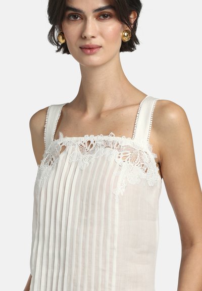 Femme portant un haut blanc sans manches plissé avec un détail en dentelle florale le long de l'encolure et des boucles d'oreilles rondes dorées.