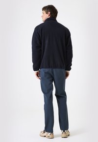 Chaqueta de forro polar azul oscuro con un alto cuello, puños elásticos y un corte relajado, combinada con pantalones oscuros y zapatillas de color claro.
