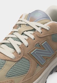 Gros plan sur une sneaker New Balance beige et vert clair avec mesh texturé, superpositions en daim, lacets blancs et grand logo "N" sur le côté.