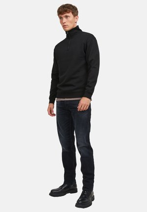 Man met een zwarte half-rits trui, donkere spijkerbroek en zwarte veterboots, staand tegen een effen witte achtergrond.