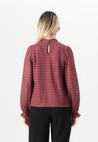 Vrouw met lang blond haar draagt een gebloemde bordeauxrode blouse met lange mouwen en een keyhole knoopsluiting, gecombineerd met zwarte broek, en kijkt weg.