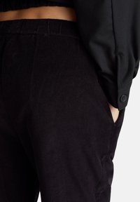 Detailní záběr osoby, která má na sobě černé manšestrové kalhoty s elastickým pasem a černou košili s dlouhým rukávem a knoflíkovým manžetou, ruka v kapse.