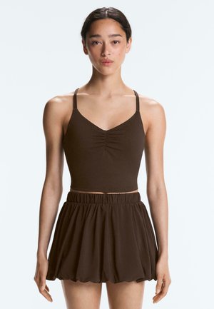 Jonge vrouw draagt een bruin geplooid cropped top met dunne bandjes en een bijpassende hoog getailleerde, geplooide minirok, staande tegen een effen achtergrond.