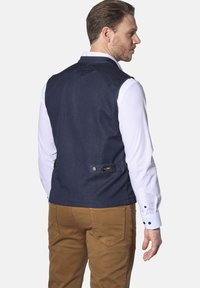 Dunkelblauer Jeanswesten mit glatter Textur, maßgeschneiderter Passform und verstellbaren Seitenriemen mit Knöpfen. Über einem weißen Hemd getragen.