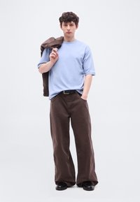 Νεαρός με σγουρά μαλλιά φοράει ανοιχτό μπλε μπλουζάκι, φαρδιά καφέ παντελόνια, μαύρα παπούτσια, κρατώντας καφέ σακάκι στον ώμο, στέκεται σε λευκό φόντο.