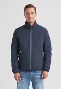 Marineblaue Jacke mit Reißverschluss, hohem Kragen und zwei seitlichen Taschen. Verfügt über einen kontrastierenden hellblauen Reißverschluss und ausgeprägte Schulternahtnähte.