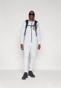 Lys grå hettegenser med glidelås og snor, kombinert med matchende joggebukser. Svart ryggsekk med gule og oransje detaljer. Svart sneakers.