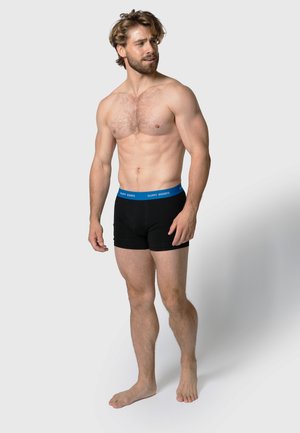 Zwarte katoenen boxershorts met een blauwe elastische tailleband, met de merknaam "HAPPY SHORTS" aan de voorkant. Model met een lichte huid en kort haar.