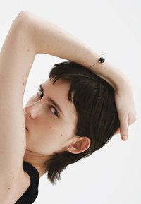 Pulsera de piedra negra en la muñeca, banda dorada delgada y diseño minimalista. Primer plano de una persona con cabello corto, que muestra joyas en las orejas.