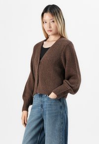 Kvinne med langt blondt hår iført en brun strikket cardigan over en svart topp og vide blå jeans, stående med den ene hånden i lommen.