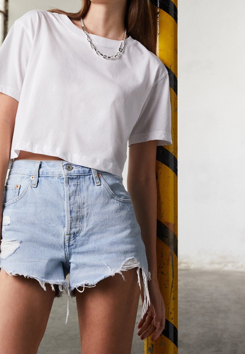 Personne portant un t-shirt blanc court, un short en jean clair déchiré et un collier chaîne en argent, appuyée contre un poteau rayé jaune et noir.