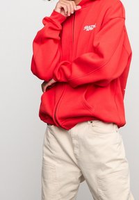 Personne portant un sweat à capuche zippé rouge vif avec le texte "MULTI APP" et un pantalon cargo crème, posant avec la main près du visage et de la hanche.