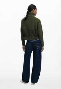 Giacca a maglia verde con un pannello verde scuro nella parte inferiore, abbinata a jeans blu scuro a gamba larga con cuciture decorative e un leggero luccichio.