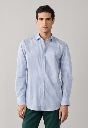 Hombre con cabello oscuro que lleva una camisa de manga larga abotonada con rayas blancas y azul claro, y pantalones verde oscuro, de pie frente a un fondo liso.