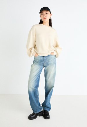 Felpa oversized crema, jeans a gamba larga blu chiaro e scarpe nere a punta brillante. Tessuto liscio, vestibilità rilassata e finitura in denim sbiadita.