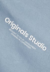 Tessuto azzurro chiaro con una superficie liscia, con testo ricamato bianco: "Originals Studio", accompagnato da ulteriore testo sottostante.