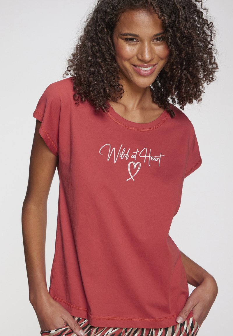 Červené bavlnené tričko s krátkymi rukávmi, na ktorom je text "Wild at Heart" v bielej písanej tlači. Mäkká textúra a voľný strih.