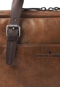 Braune Lederhandtasche mit einem dunkelbraunen Riemen und einer silbernen Metall Schnalle; geprägtes Logo an der Seite; glatte Oberfläche mit minimalen Nähten.