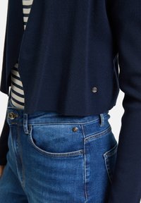 Cardigan a maglione blu navy con un design corto, abbinato a jeans in denim blu con cuciture a vista e dettagli in metallo sui bottoni.
