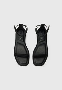 Čierne sandále s plochou podrážkou, gumovými a textilnými popruhmi, otvoreným dizajnom päty a textúrovaným povrchom. Logo "DKNY" viditeľné na vnútorniej strane.