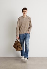 Beige langermet polo skjorte, blå falmede jeans, og hvite joggesko. Modellen bærer en brun semsket lærveske med lommer. Nøytral bakgrunn.
