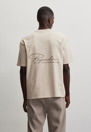 Homme aux cheveux courts portant un T-shirt beige avec le texte « Pegador All Rights Reserved » au dos et un pantalon côtelé beige clair.